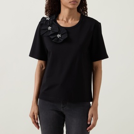 Model wearing Joseph Ribkoff black top with floral corsage and crystal embellishments at shoulder, paired with dark grey jeans.