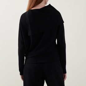 Model wearing Joseph Ribkoff black long-sleeve top, rear view showing asymmetric neckline detail and relaxed fit, styled with black trousers.