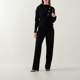 Model wearing Joseph Ribkoff black top with draped overlay and gold buttons, styled with matching black wide-leg trousers and strappy heels.