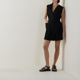Model wearing Joseph Ribkoff black sleeveless wrap-style playsuit with tie waist, styled with black chunky strap sandals