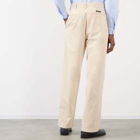 Model wearing Prada sand cotton trousers, rear view, styled with a light blue shirt, relaxed straight-leg fit with back patch label detail.