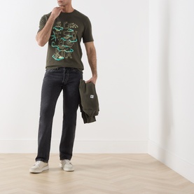 Model wearing Scotch & Soda Forest Night green graphic cotton T-shirt, styled with dark jeans and white trainers, holding a jacket.