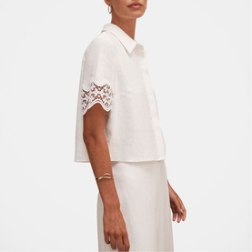 Model wearing white cropped short-sleeve shirt with lace trim sleeves, styled with matching white midi skirt and gold bracelet.