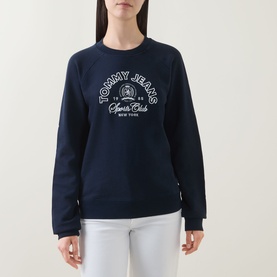 Model wearing Tommy Jeans Dark Night Navy cotton sweatshirt with varsity Sports Club graphic print, styled with white trousers.