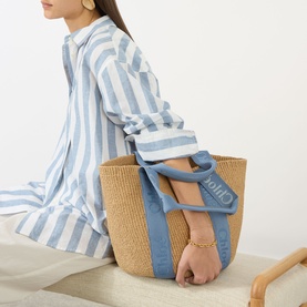 Model carrying Chlo&eacute; Woody Large Basket Tote Bag in Ashes Blue, woven paper with logo tape handles, styled with a blue striped shirt dress.
