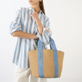 Model carrying Chlo&eacute; Woody Large Basket Tote Bag in Ashes Blue, natural woven body with blue logo tape handles, styled with striped shirt