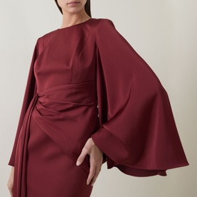 Model wearing Maisonel Draped Pencil Dress in brick, featuring structured shoulders, cape-style long sleeves and ruched waist detail.
