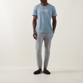 Model wearing Ralph Lauren Script Logo Jersey Sleep Shirt in Cool Grey Blue with grey joggers and navy slippers.