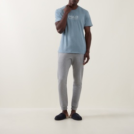 Model wearing Ralph Lauren Script Logo Jersey Sleep Shirt in Cool Grey Blue with grey joggers and navy slippers.
