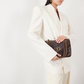 Model carrying Valentino Djuna Medium Suede Shoulder Bag in Oak Brown with gold-tone chain strap and V-logo hardware, styled with white blazer