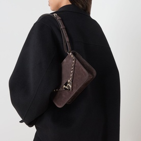 Model carrying Valentino Rockstud Small Suede Shoulder Bag in oak brown with gold-tone hardware and stud embellishment, styled over black coat.
