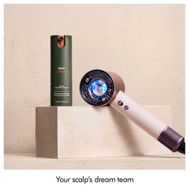 Dyson hair dryer in pink and copper tones styled with a green Dyson hair serum bottle on a stone plinth