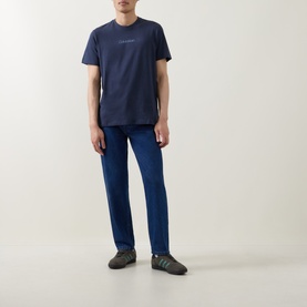 Model wearing Calvin Klein Graphic Logo T-Shirt in Blue Nights, styled with navy trousers and grey trainers.
