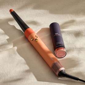 Dyson hair styling tool in coral orange with gold accents, shown with detachable barrel attachment and purple storage cap