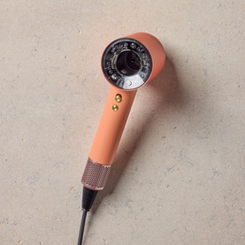 Dyson Supersonic hair dryer in coral orange with rose gold handle, shot from above on neutral stone surface