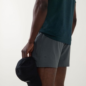 Model wearing Lululemon Pace Breaker Shorts 5" in Onyx Grey, styled with a teal vest top, holding a black cap.