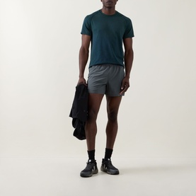 Model wearing Lululemon Pace Breaker Shorts 5" in Onyx Grey, styled with a teal T-shirt and dark trainers, holding a jacket.
