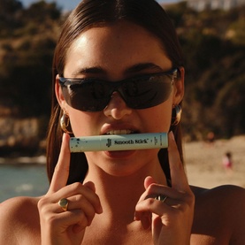 Model with The Smooth Company Smooth Stick&trade; Island Edition held to lips, wearing black sunglasses and gold rings, beach setting