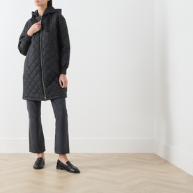 Model wearing Marc O Polo black quilted hooded coat with zip fastening, styled with dark cropped trousers and black loafers.