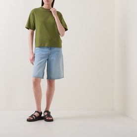 Model wearing Marc O Polo Solid Relaxed T-Shirt in Shaded Fern, styled with light blue denim shorts and black chunky sandals.