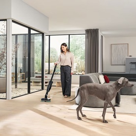 Model using Miele Triflex HX3 Vacuum Cleaner Aqua on hardwood floor in bright living room, with a large grey dog nearby.