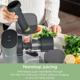 Ninja cold press juicer in grey with pusher tool, shown in use with fresh vegetables and citrus on a kitchen counter.