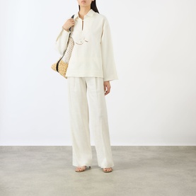 Model wearing Victoria Beckham white linen-look top with wide sleeves and zip neck, styled with matching wide-leg trousers and straw bag.