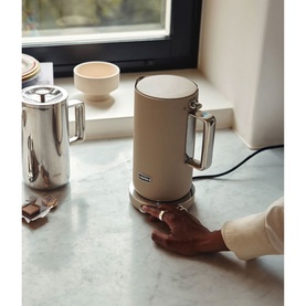 Aarke Kettle Sand in cream with brushed metal base and handle, shown on marble surface beside a chrome blender, hand pressing base button