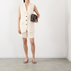 Model wearing Anine Bing cream gilet with matching shorts, carrying a dark brown mini bucket bag, styled with tan flip flops.