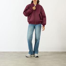Model wearing Anine Bing Leon Bomber Jacket in burgundy with zip-through fastening, styled with light blue jeans and grey trainers.