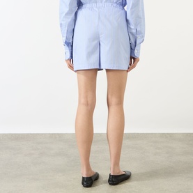 Model wearing Anine Bing Ramy Striped Shorts in washed blue with elasticated waistband, styled with matching shirt and black flats. Rear view.