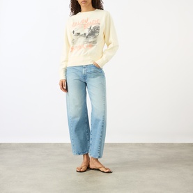 Model wearing Anine Bing Washed Buttercream graphic sweat top, styled with light blue wide-leg jeans and tan flat sandals.