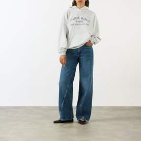 Model wearing Anine Bing grey logo hoodie sweatshirt with wide-leg blue jeans and black flat shoes.
