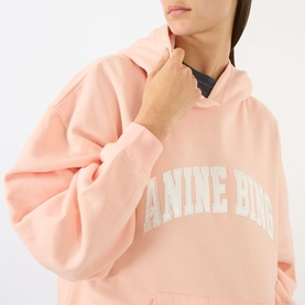 Model wearing Anine Bing blush hoodie sweatshirt with oversized logo print, adjusting hood drawstring