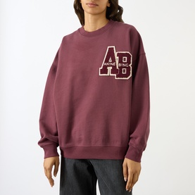 Model wearing Anine Bing burgundy cotton sweatshirt with varsity AB logo patch, styled with grey wide-leg trousers.
