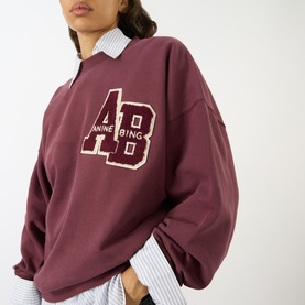 Model wearing Anine Bing burgundy cotton sweat top with varsity AB logo, styled over a striped collared shirt.