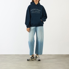 Model wearing Anine Bing navy cotton hoodie with logo print, styled with light wash wide-leg jeans and white trainers.