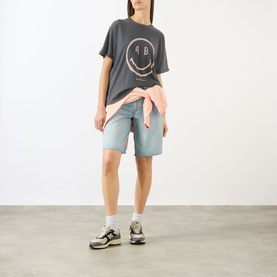 Model wearing Anine Bing washed grey graphic smiley face T-shirt, styled with light blue denim shorts and a pink tied shirt at waist.