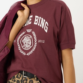 Model wearing Anine Bing burgundy cotton T-shirt with collegiate crest graphic, styled with leopard-print skirt and burgundy jacket.