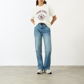 Model wearing Anine Bing ecru cotton T-shirt with burgundy logo graphic, styled with mid-blue straight-leg jeans and silver trainers.