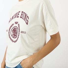 Model wearing Anine Bing ecru cotton T-shirt with burgundy collegiate crest graphic, styled with blue denim jeans.