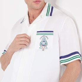 Model wearing Casablanca Hotel Emblem Silk Shirt in white with green and navy stripe trims, camp collar, and chest patch pocket with graphic.