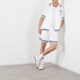 Model wearing Casablanca Hotel Emblem Silk Shirt in white with stripe trims, styled with matching white shorts and white trainers.