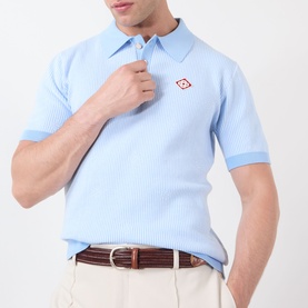 Model wearing Casablanca Pinstripe Logo Polo Shirt in light blue with diamond logo at chest, styled with cream trousers and brown belt.