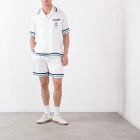 Model wearing Casablanca Hotel Emblem Silk Shorts in white with navy stripe trims, styled with matching short-sleeve shirt and white trainers.