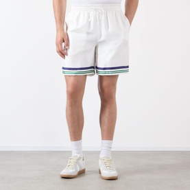 Model wearing Casablanca Hotel Emblem Silk Shorts in white with blue and green stripe hem trim, styled with white socks and cream trainers.