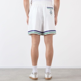 Model wearing Casablanca Hotel Emblem Silk Shorts in white with navy and green stripe hem trim, rear emblem patch pocket, paired with white trainers.