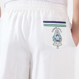 Model wearing Casablanca Hotel Emblem Silk Shorts in white, showing rear patch pocket with embroidered hotel emblem and navy stripe trim detail.