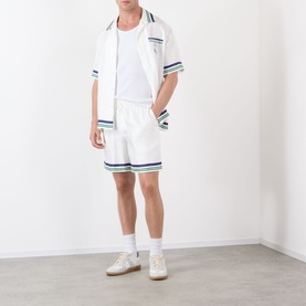 Model wearing Casablanca Hotel Emblem Silk Shorts in white with blue and green stripe trims, styled with matching shirt and white trainers.