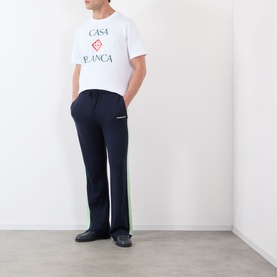 Model wearing Casablanca Diamond Logo T-Shirt in white with stacked logo graphic, styled with navy side-stripe trousers and dark loafers.